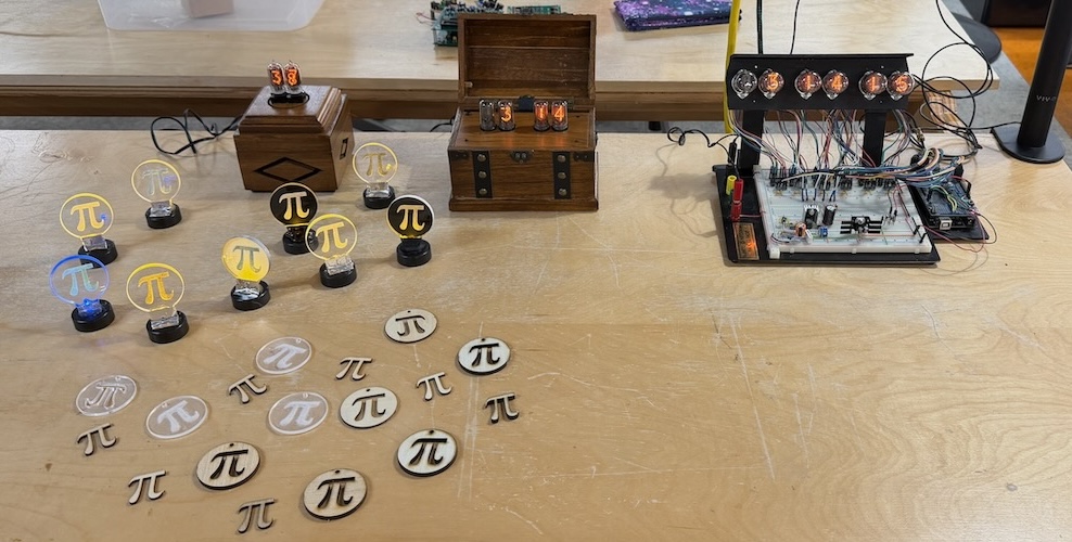 A workbench covered in Π Day creations — laser-cut π symbols in various sizes, LED-lit acrylic π signs on small bases, a breadboarded Nixie tube driver circuit, and wooden boxes with Nixie tubes displaying the digits of Π — well, actually — the current time